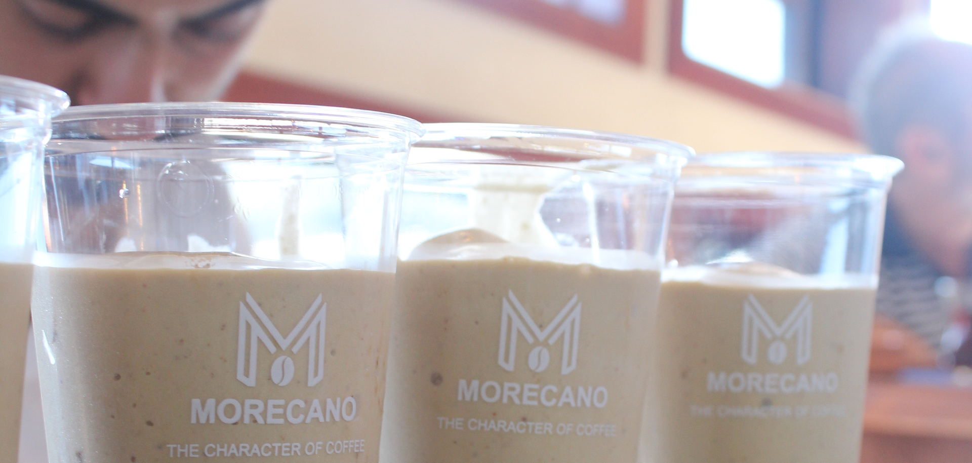 Tezgah üstünde hazırlanmış üç Morecano bardağı; arkada servis eden barista